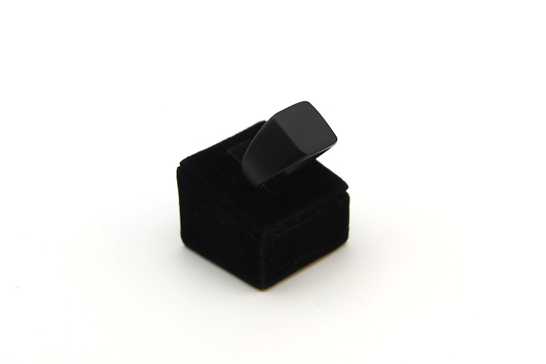 May include: A black ring with a square top is displayed on a black velvet ring holder. The ring has a simple, modern design. The ring holder is a small, square block.
