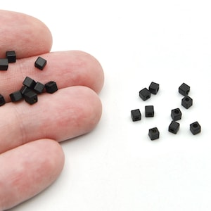 May include: Small, black, square beads with a hole through the centre are scattered on a white surface. Some beads are held in a hand. These beads are likely used for jewellery making or other crafts.