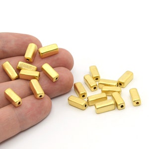 May include: Gold-colored, rectangular beads with rounded edges. The beads are scattered on a white surface, with some held in a hand. Each bead has a small hole at each end, suitable for stringing. These beads are ideal for jewelry making.