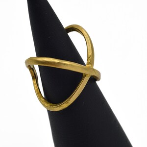 May include: A gold-colored ring with an open, geometric design. The ring features a curved band and an upper element that crosses over the band. The ring is displayed on a black cone-shaped stand against a white background.