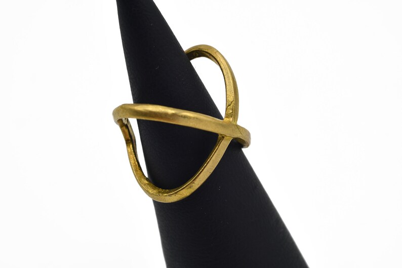 May include: A gold-colored ring with an open, geometric design. The ring features a curved band and an upper element that crosses over the band. The ring is displayed on a black cone-shaped stand against a white background.