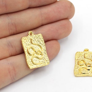 May include: Two gold-coloured rectangular pendants with a textured surface and a snake design. Each pendant has a small loop at the top for attaching to a chain or other jewellery component. The pendants are shown against a white background.