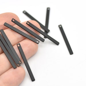 May include: A collection of matte black metal bar charms with a small hole at one end. These rectangular charms are scattered on a white surface. The charms are approximately 1 inch long.
