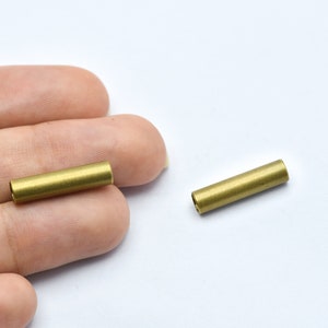 May include: Two small, cylindrical brass tubes are shown against a white background. The tubes are golden in color and appear to be hollow. The tubes are approximately 0.75 inches long and 0.25 inches in diameter. They are likely components for jewelry or crafts.