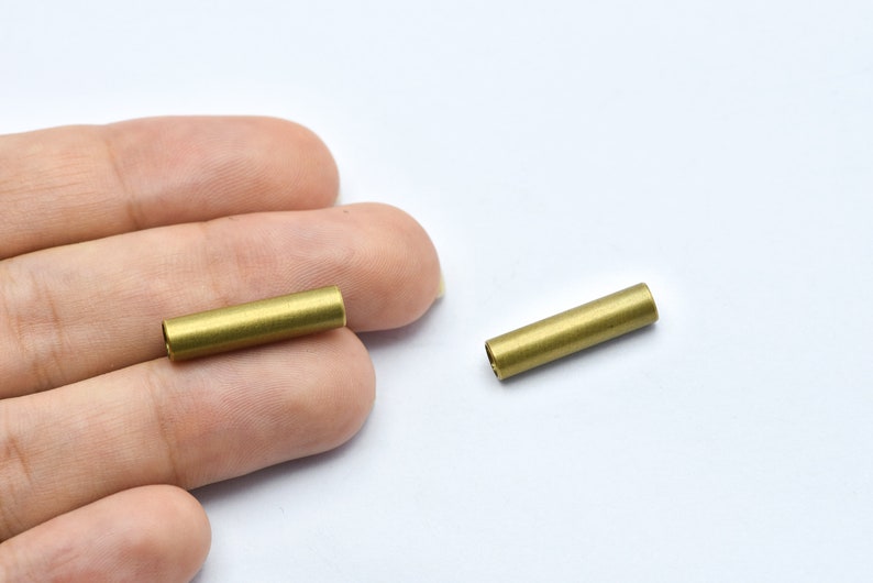 May include: Two small, cylindrical brass tubes are shown against a white background. The tubes are golden in color and appear to be hollow. The tubes are approximately 0.75 inches long and 0.25 inches in diameter. They are likely components for jewelry or crafts.