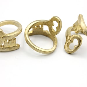 May include: Three gold-colored key-shaped rings are displayed against a white background. The rings have a keyhole design and a textured finish. The keys are in different positions, showcasing the design from various angles. The rings are likely jewelry.