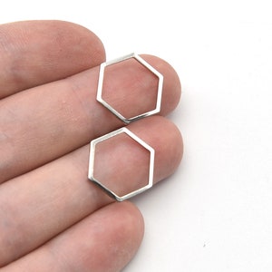 May include: Two small, silver-colored, open-frame hexagon-shaped charms. The charms are held in a person's hand against a white background. The charms are likely for jewelry making or crafting.