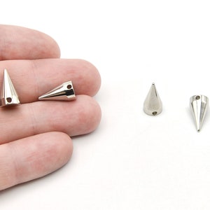 May include: Close-up of several silver-colored metal cone-shaped studs. Each stud has a pointed tip and a small hole near the base. The studs are displayed against a white background.