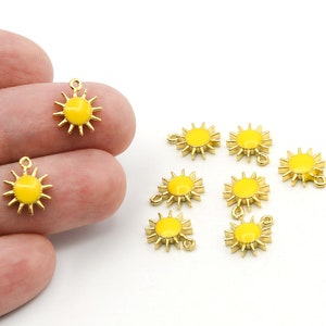 Puede incluir: Amuletos de sol dorados con centros de esmalte amarillo. Los amuletos de sol tienen un lazo en la parte superior para sujetarlos a las joyas. Los amuletos son pequeños, con un centro amarillo brillante y rayos dorados.