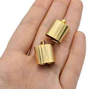 May include: Two gold-coloured, cylindrical jewellery components with small loops on top. The components are held in a hand against a white background. The cylinders have a shiny, reflective surface.