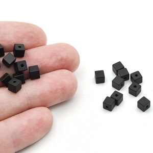 May include: Small, black, square beads with a central hole, scattered on a white background and in a hand. These beads are suitable for jewellery making and various craft projects. Each bead is approximately 5 mm.