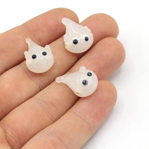 以下が含まれることがあります： 黒い目を持つ、半透明のゴースト型のビーズが3つ。白い背景に、手でビーズを持っています。これらのビーズは、ジュエリー作りやその他のクラフトプロジェクトに使用される可能性があります。