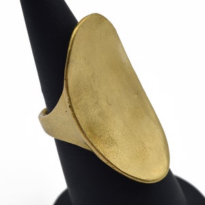 May include: A large, gold-colored ring with an elongated oval face. The ring is displayed on a black stand against a white background. The ring's surface has a slightly textured appearance, suggesting a brushed metal finish.