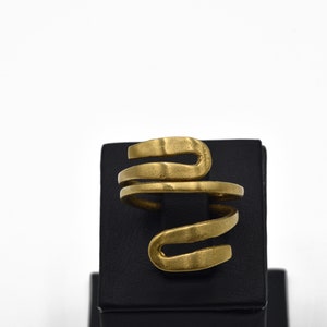 May include: A gold-colored ring with a spiral design. The ring is made of a textured metal and sits on a black display stand. The ring's design resembles a snake or a coiled spring.