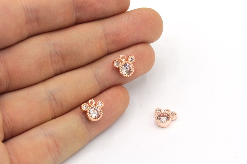 May include: Rose gold-colored charms in the shape of a mouse head, each with a clear, round center stone and three smaller stones for the ears. The charms are small, delicate, and suitable for jewelry making.