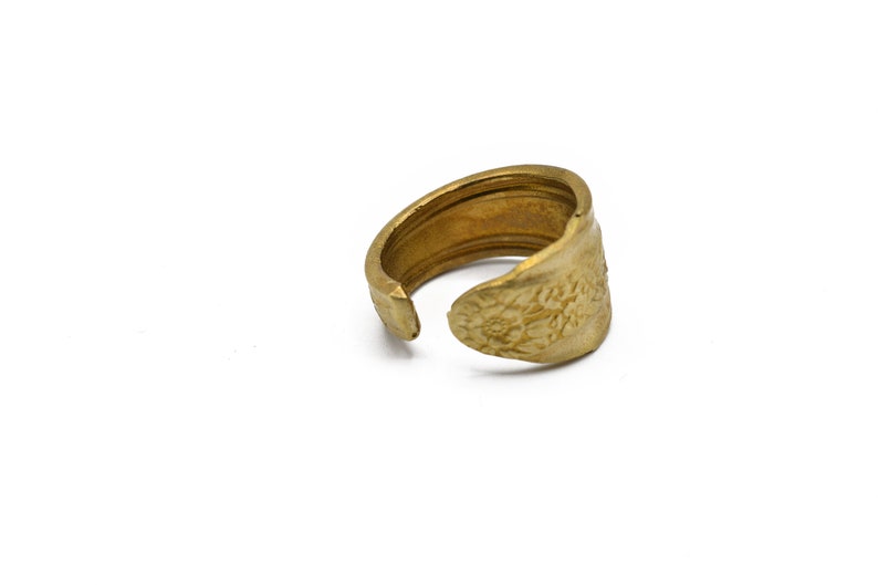 May include: A handcrafted brass ring with a floral design on the face. The adjustable ring is made from a repurposed spoon, showcasing a unique, vintage aesthetic. The ring is a warm gold color.