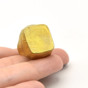 Puede incluir: Un anillo dorado con una banda texturizada y una parte superior cuadrada. El anillo se sostiene en la mano de una persona sobre un fondo blanco. La parte superior cuadrada del anillo tiene una superficie lisa.