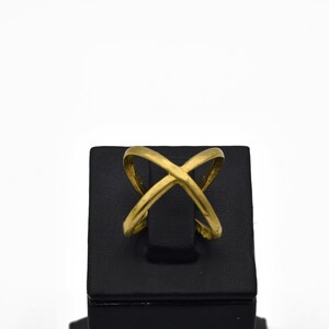 May include: A gold-colored ring with an overlapping X design. The ring is displayed on a black stand against a white background. The ring's design features two curved bands that intersect.