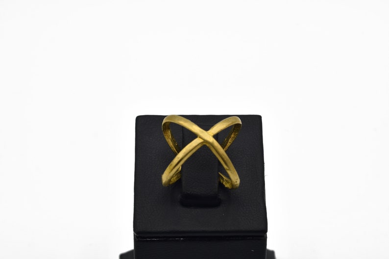 May include: A gold-colored ring with an overlapping X design. The ring is displayed on a black stand against a white background. The ring's design features two curved bands that intersect.