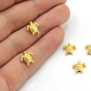 May include: A collection of small, gold-coloured turtle charms are shown. Each charm has a textured shell and flippers. These miniature turtle figurines are likely for jewellery making or crafting, and are set against a white background.