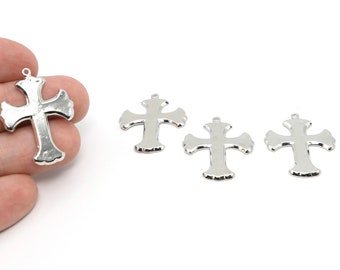 May include: Silver-colored cross pendants with a decorative, scalloped edge. The crosses have a loop at the top for stringing. The image shows four crosses, one held by a hand, against a white background.