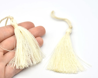 May include: Two cream-colored tassels with a looped cord at the top. The tassels are made of many thin strands, and the background is white. These decorative tassels can be used for various craft projects.