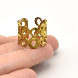 May include: A gold-colored ring with a repeating circular design. The ring is made of metal and has an open, adjustable design. The ring is shown on a person's hand against a white background. The ring's design is made of connected circles.