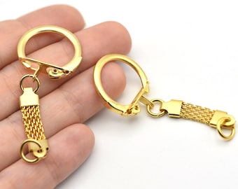 Puede incluir: Dos llaveros dorados con un diseño de cadena de malla. Cada llavero presenta un cierre circular grande y un conector rectangular. La cadena de malla cuelga del conector, terminando en un pequeño anillo.