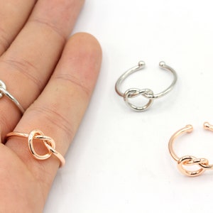 May include: Four adjustable knot rings in silver and rose gold tones are displayed against a white background. The rings feature a knot design at the center, with an open band for adjustability. The rings are a simple, elegant jewelry item.
