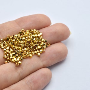 以下が含まれることがあります： 小さな金色のファセットビーズのコレクション。ビーズは白い背景に対して手で持たれています。ビーズは金属的な光沢と幾何学的な形状をしており、ジュエリー作りや工芸品に使用されていることを示唆しています。
