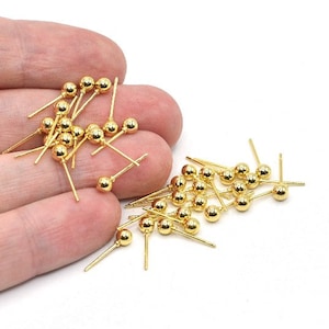 May include: A collection of small, gold-colored jewelry findings. Each finding has a small, round bead at the top of a thin, straight post. The findings are scattered on a white surface.