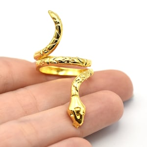May include: A gold-colored snake ring with a coiled design. The ring features a detailed snake head and body, with textured patterns. The ring is shown on a hand against a white background.