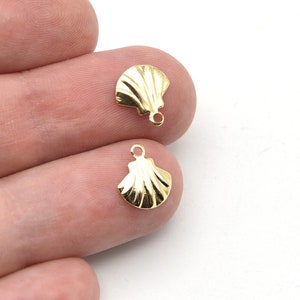 May include: Two small, gold-colored seashell charms. Each charm has a detailed, ridged surface and a small loop at the top for attaching to jewelry. The charms are displayed against a white background.