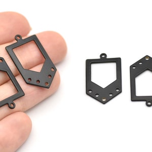 Könnte beinhalten: Schwarze Metallanhänger mit geometrischen Formen. Die Anhänger haben einen rechteckigen Rahmen mit einem ausgeschnittenen Design und kleinen Löchern zur Befestigung. Die Anhänger werden auf weißem Hintergrund gezeigt.