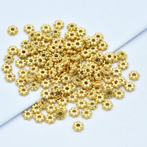 Peut inclure: Un ensemble de petites perles dor&eacute;es en forme de fleur, dispers&eacute;es sur une surface blanche. Chaque perle pr&eacute;sente un trou central et de multiples protub&eacute;rances en forme de p&eacute;tales, cr&eacute;ant un aspect d&eacute;licat et d&eacute;coratif. Ces perles sont probablement utilis&eacute;es pour la fabrication de bijoux ou des projets d'artisanat.