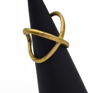 May include: A gold-colored ring with an open, geometric design. The ring features an overlapping X shape with a curved top element. The ring is displayed on a black stand against a white background.