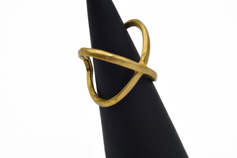 May include: A gold-colored ring with an open, geometric design. The ring features an overlapping X shape with a curved top element. The ring is displayed on a black stand against a white background.