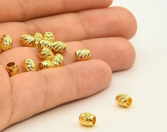 May include: A collection of small, gold-colored beads with a textured, ridged surface. The beads are cylindrical with a hole running through the center. The beads are scattered in a person's hand against a white background. These beads are suitable for jewelry making.