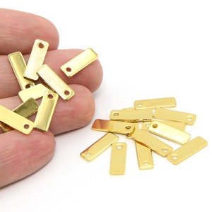 May include: A collection of small, rectangular gold-colored metal tags with a single hole at one end. The tags are shown against a white background, with some held in a hand.