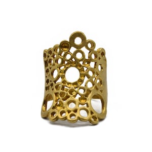 Op de afbeelding: Een goudkleurige ring met een open ontwerp met cirkels van verschillende grootte. De ring heeft een brede band en een decoratieve bovenkant, wat een unieke en artistieke uitstraling creëert. De sieraden staan tegen een witte achtergrond.