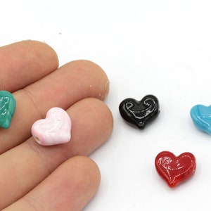 May include: A hand holds five heart-shaped beads in various colors: green, pink, black, blue, and red. The beads are glossy and appear to be made of glass or a similar material. The background is white.