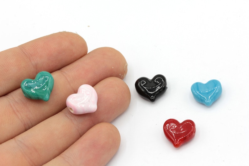 May include: A hand holds five heart-shaped beads in various colors: green, pink, black, blue, and red. The beads are glossy and appear to be made of glass or a similar material. The background is white.