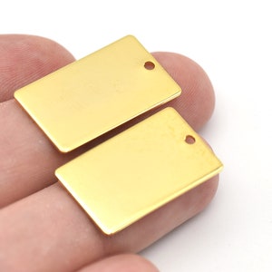 Pode incluir: Duas etiquetas retangulares de metal dourado, com cantos arredondados e um pequeno orifício na parte superior. As etiquetas são lisas e brilhantes, e são seguradas na mão contra um fundo branco. Estas etiquetas são provavelmente usadas para fazer joias ou artesanato.