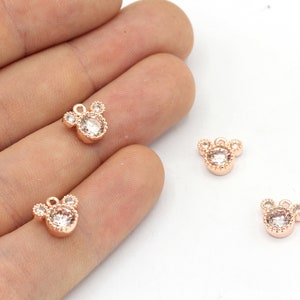 May include: Rose gold-colored charms in the shape of a mouse head, each with a central clear crystal and three smaller crystals. The charms have a small loop at the top for attaching to jewelry. The charms are shown on a light background.
