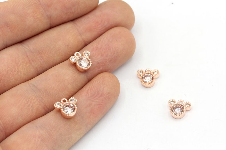 May include: Rose gold-colored charms in the shape of a mouse head, each with a central clear crystal and three smaller crystals. The charms have a small loop at the top for attaching to jewelry. The charms are shown on a light background.