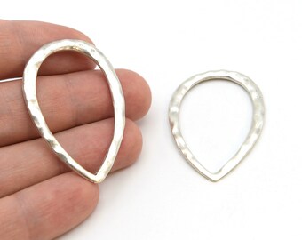 Puede incluir: Dos componentes de joyería plateados en forma de lágrima. El metal tiene una textura martillada y la forma de lágrima está abierta en el centro. Uno se sostiene en una mano y el otro está sobre una superficie blanca.