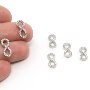 May include: Silver-toned infinity charms, each encrusted with small, clear crystals. The charms are in the shape of the number eight, and are displayed on a white background. The charms are small, and appear to be jewelry components.