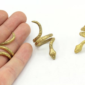 Puede incluir: Anillos de serpiente dorados. Los anillos se enrollan alrededor del dedo, con la cabeza y la cola de la serpiente levantadas. Los anillos tienen detalles grabados y se muestran sobre un fondo blanco.