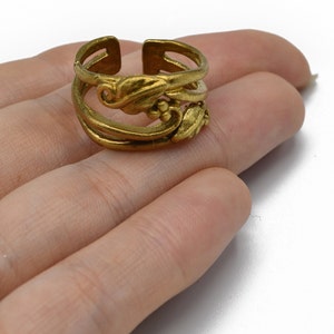 May include: A gold-colored adjustable ring with an ornate floral design. The ring features a layered band with a decorative leaf and small round accents. The ring is open at the top and is held in a hand.