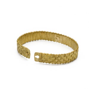 May include: A gold-colored cuff bracelet with a woven texture. The bracelet is open at one end, revealing a clasp. The design features a repeating pattern, and the overall look is vintage.
